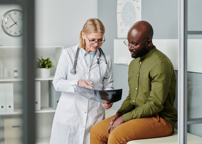 Confident mature doctor in whitecoat showing young black man x-ray image of his lungs and explaining him diagnosis in clinics